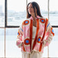 Woman wearing a colorful sweater and white pants standing in a room with large windows.