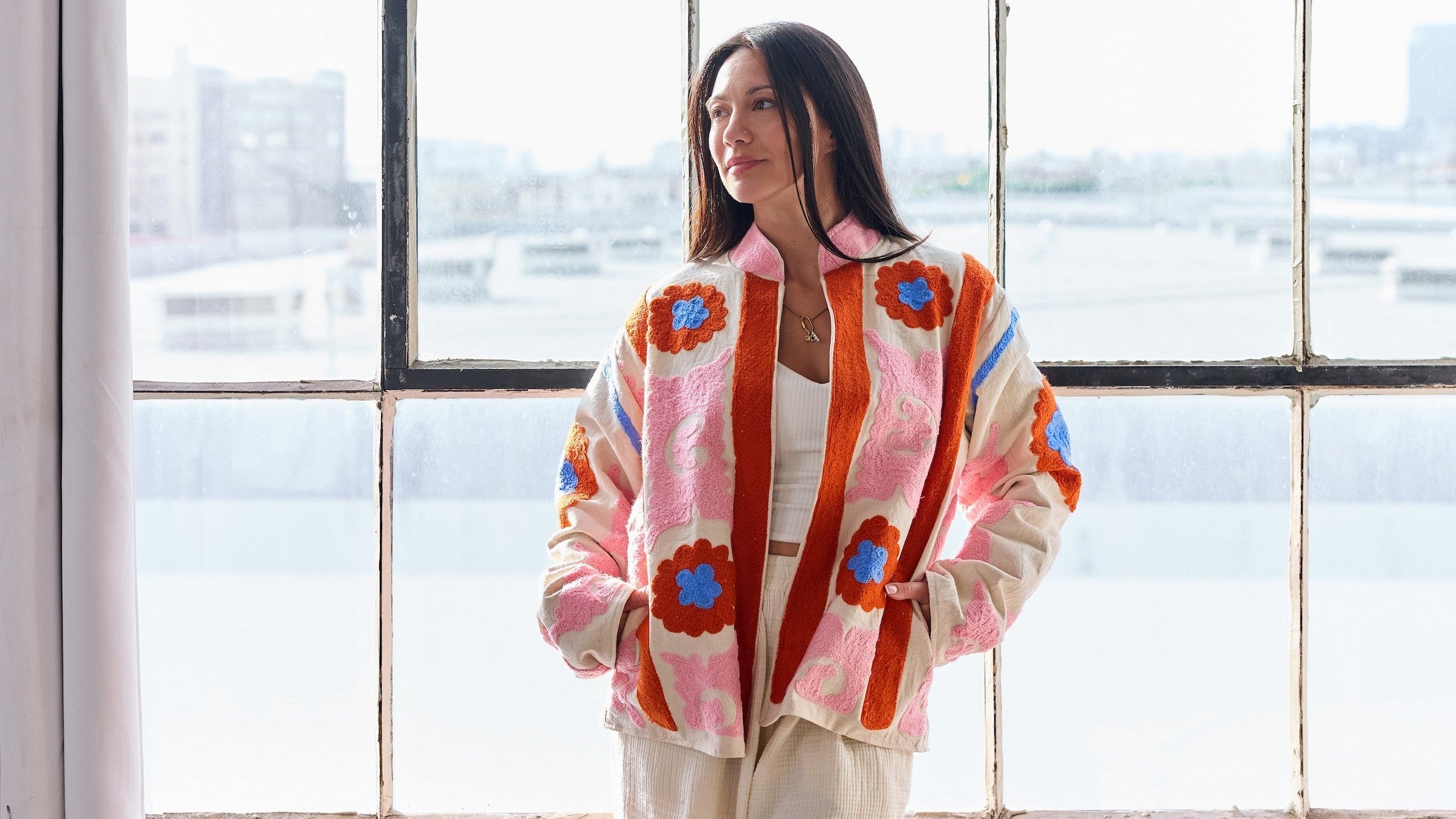 Woman wearing a colorful sweater and white pants standing in a room with large windows.